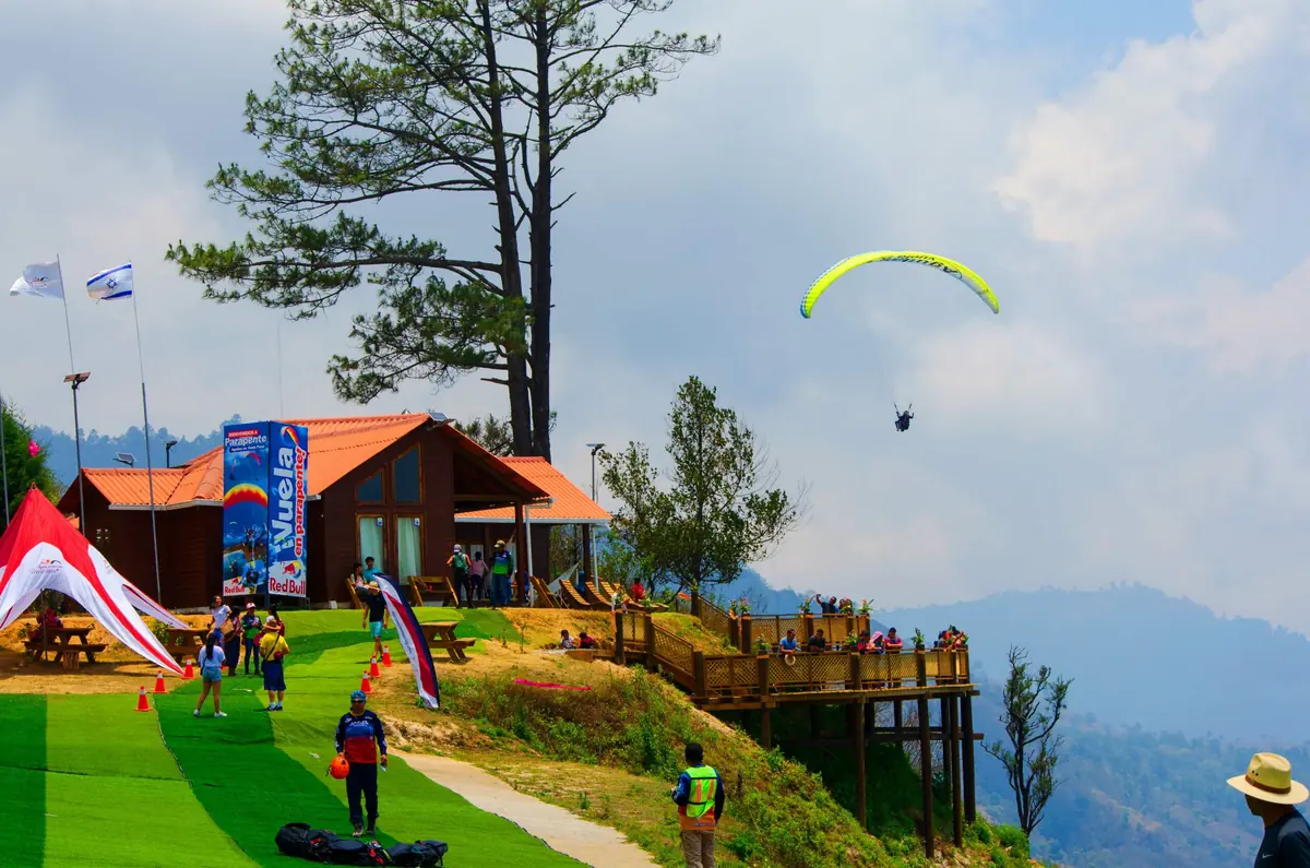 Área de despegue con visitantes, la cabaña del mirador y un parapente en el aire